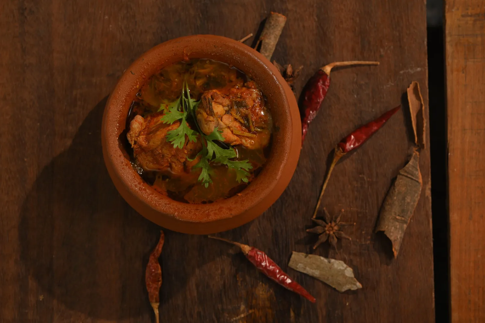 Chicken curry in a clay pot on a wooden table with dried red chillies.