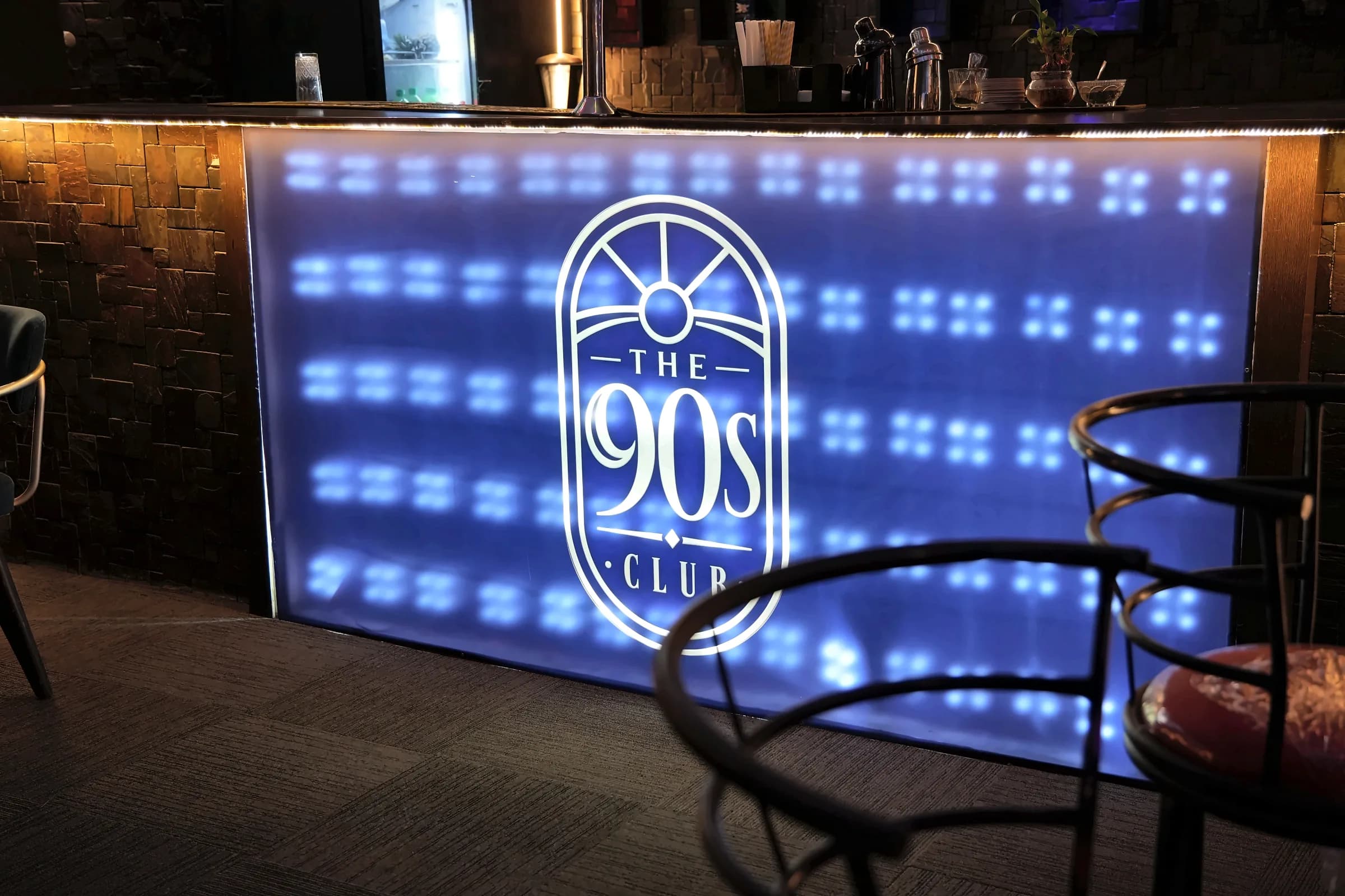 Close view of the bar front at The 90s Club, with the lit arched logo on a blue LED panel.