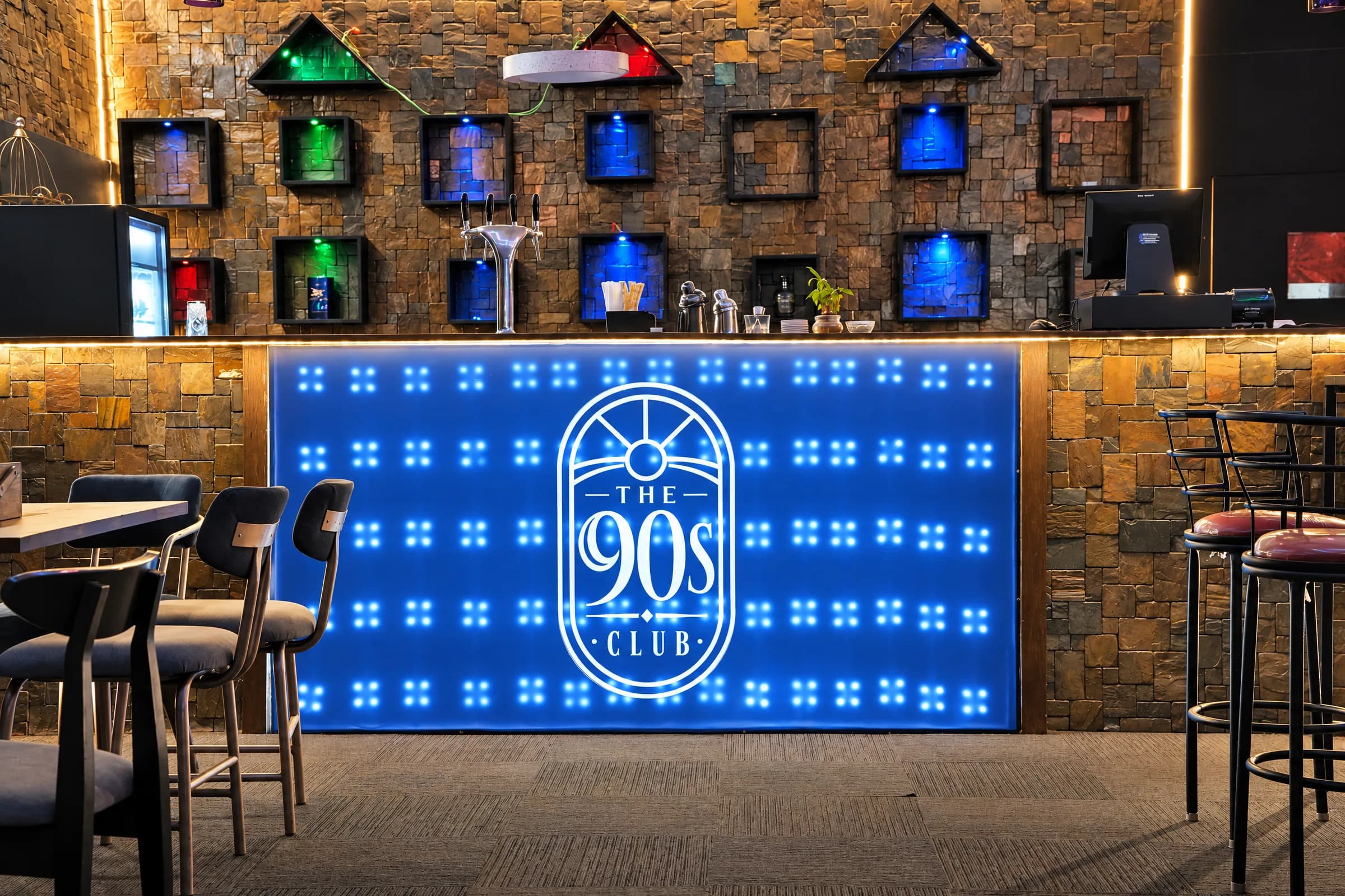 The bar counter at The 90s Club in Electronic City, with the arched 90s Club logo lit on a blue LED bar front and bar stools in front.