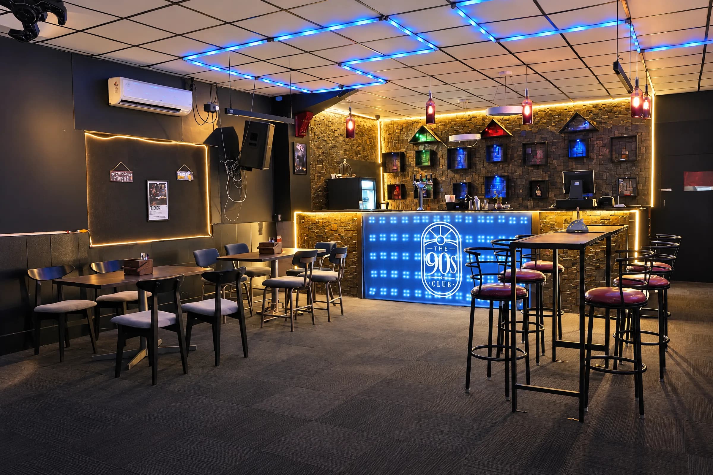 Inside The 90s Club taproom in Electronic City: blue neon lines on the ceiling, the lit logo bar and high tables with stools.